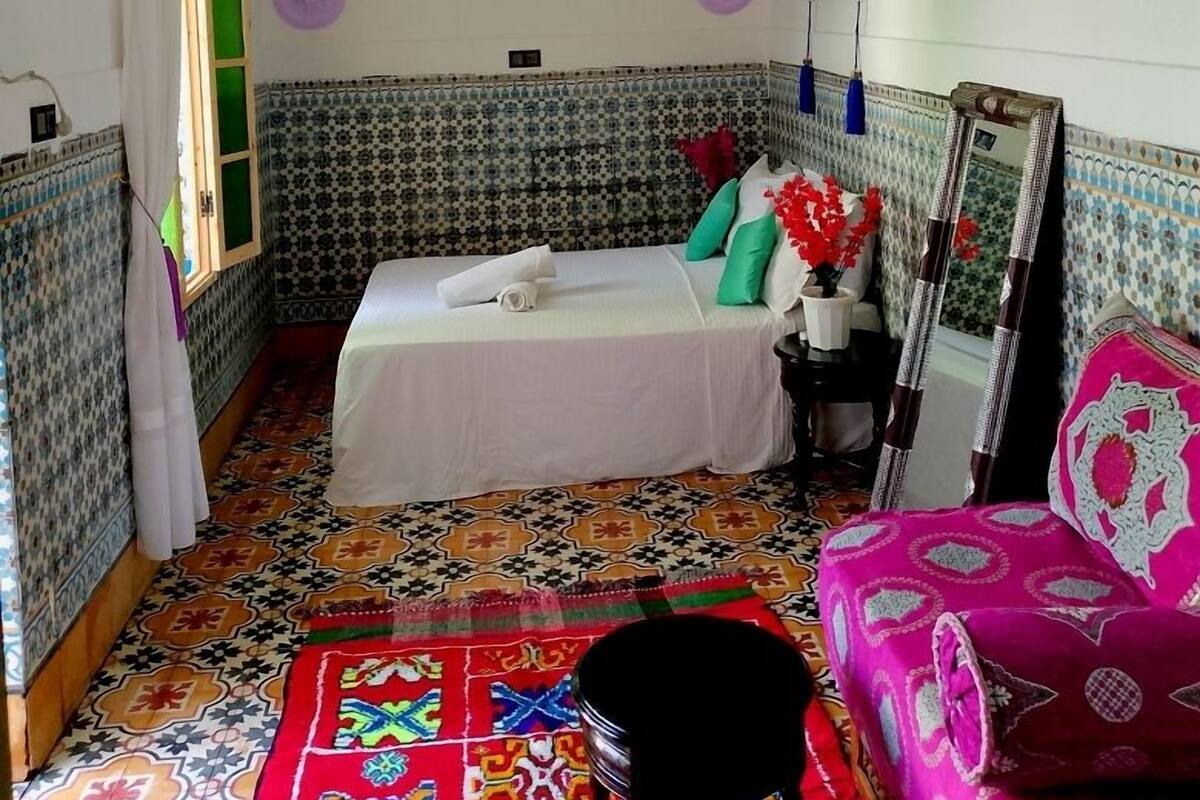 A room with a bed, a mirror, a chair, and a colorful rug.