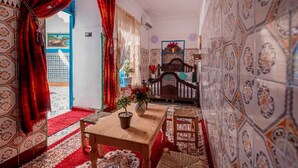 Um quarto com mesa de madeira, um banco e uma cama com cabeceira. Há uma janela com cortinas, uma parede com azulejos estampados e uma porta.