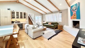 Ein großzügiger Wohnbereich im Loft-Stil mit hohen Decken, Holzbalken, einem Essbereich mit langem Tisch und Stühlen, einer Couch, einem Kamin und einer Küche mit Schränken sowie einer Treppe.