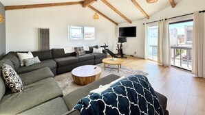 Ein geräumiges Wohnzimmer mit einer großen Couch, einem Couchtisch und einem Fernseher. Ein Balkon bietet Blick auf die Stadt.