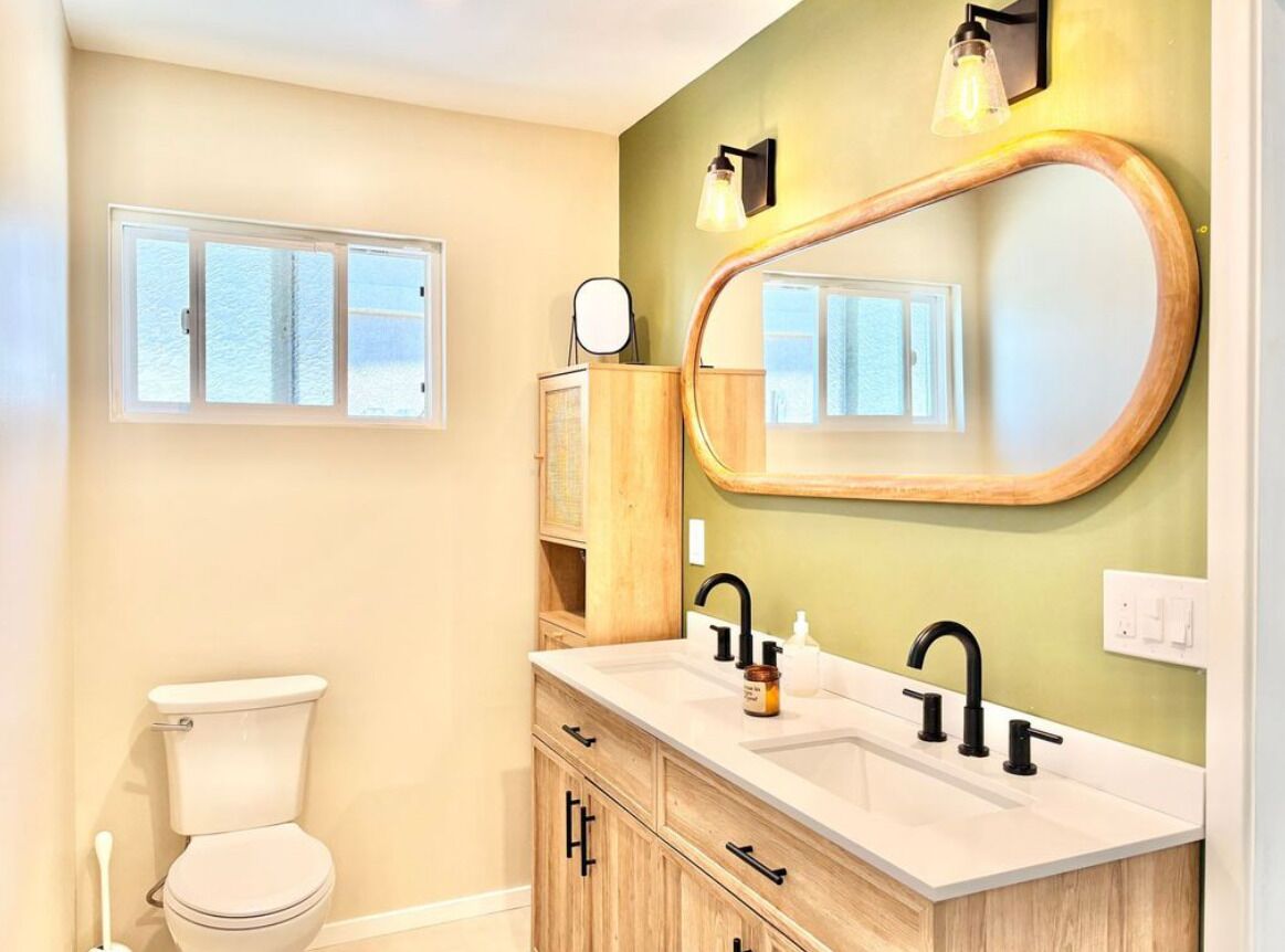 A bathroom with a toilet, a double sink vanity, a large mirror, and a window.