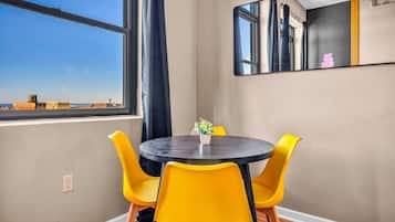 Una habitación con una mesa redonda, cuatro sillas amarillas, una planta en maceta, un ventanal con vista a edificios y un espejo en la pared.