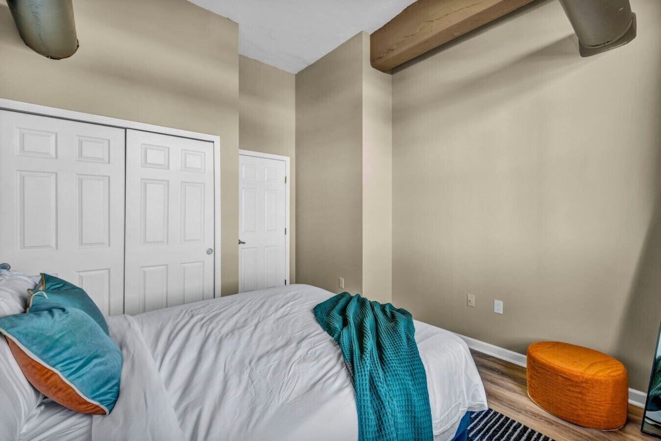 A bedroom with a bed, two doors, a wooden beam, and a small orange ottoman.
