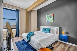 A hotel room with a large bed, a desk with a computer, a window with curtains, and a city view.