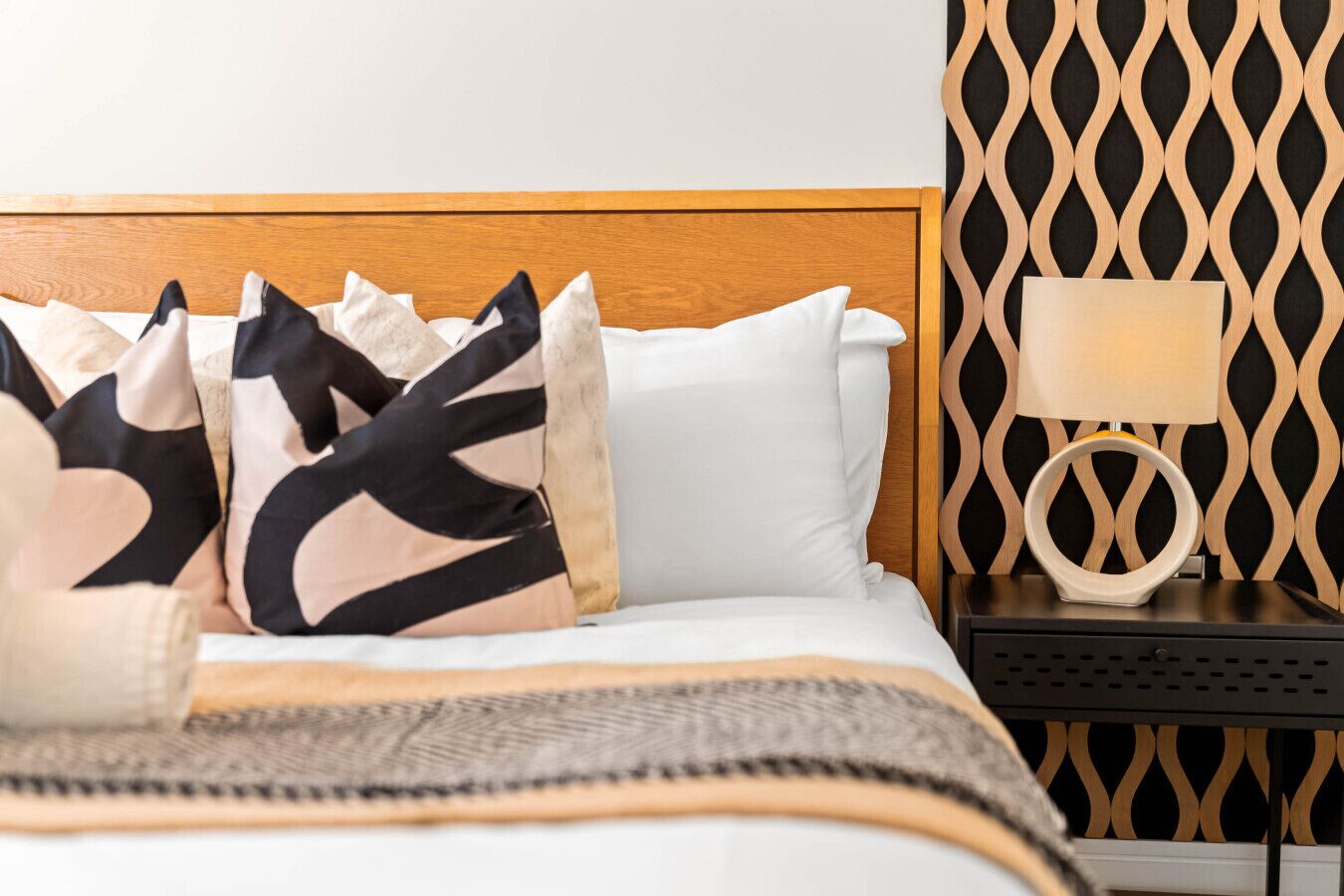 A neatly made bed with a patterned throw, a bedside table with a lamp, and a wall with a geometric pattern.