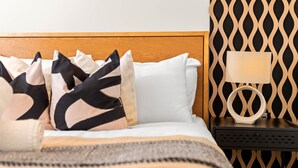 Ein ordentlich gemachtes Bett mit einer gemusterten Tagesdecke, ein Nachttisch mit einer Lampe und eine Wand mit geometrischem Muster.