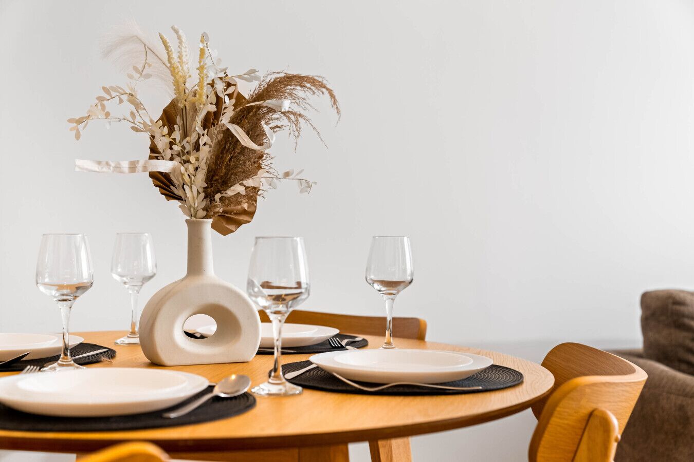 A dining table set with white plates, cutlery, and wine glasses, centered with a decorative vase of dried flowers.