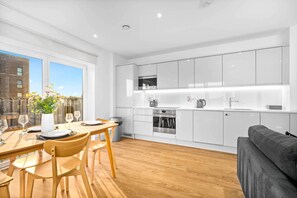 A modern kitchen with white cabinets, a dining area with wooden furniture, and a view of a building outside. - Luxury 2 Bed Apartment - Balcony - Fast WiFi - Netflix (Birmingham)
