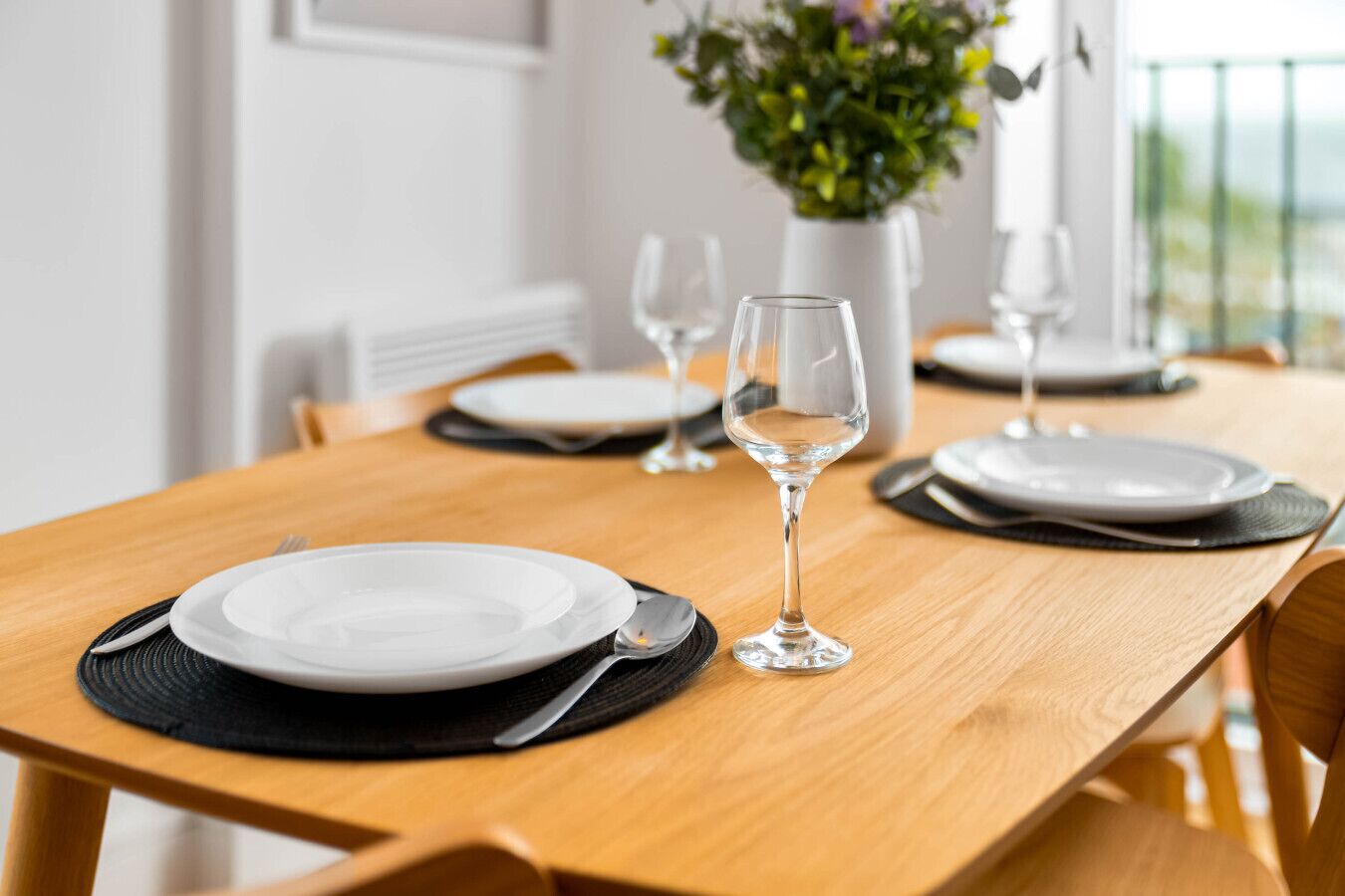 A dining table set with plates, cutlery, and wine glasses, with a potted plant in the background.