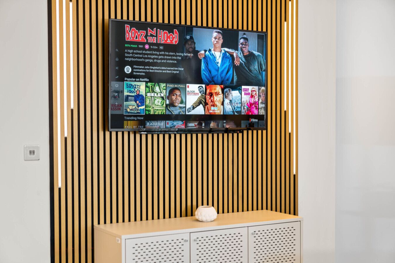 A flat-screen TV mounted on a wall with a wooden panel design, displaying a movie titled "Boyz n the Hood" and other Netflix content.