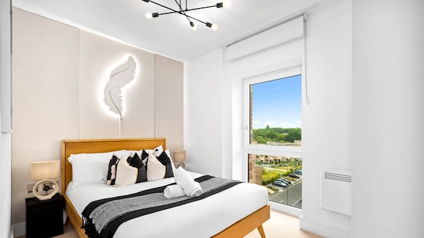 Una moderna camera d'hotel con un letto grande, un comodino con una lampada, un punto luce a parete e vista sulla città attraverso un'ampia finestra.