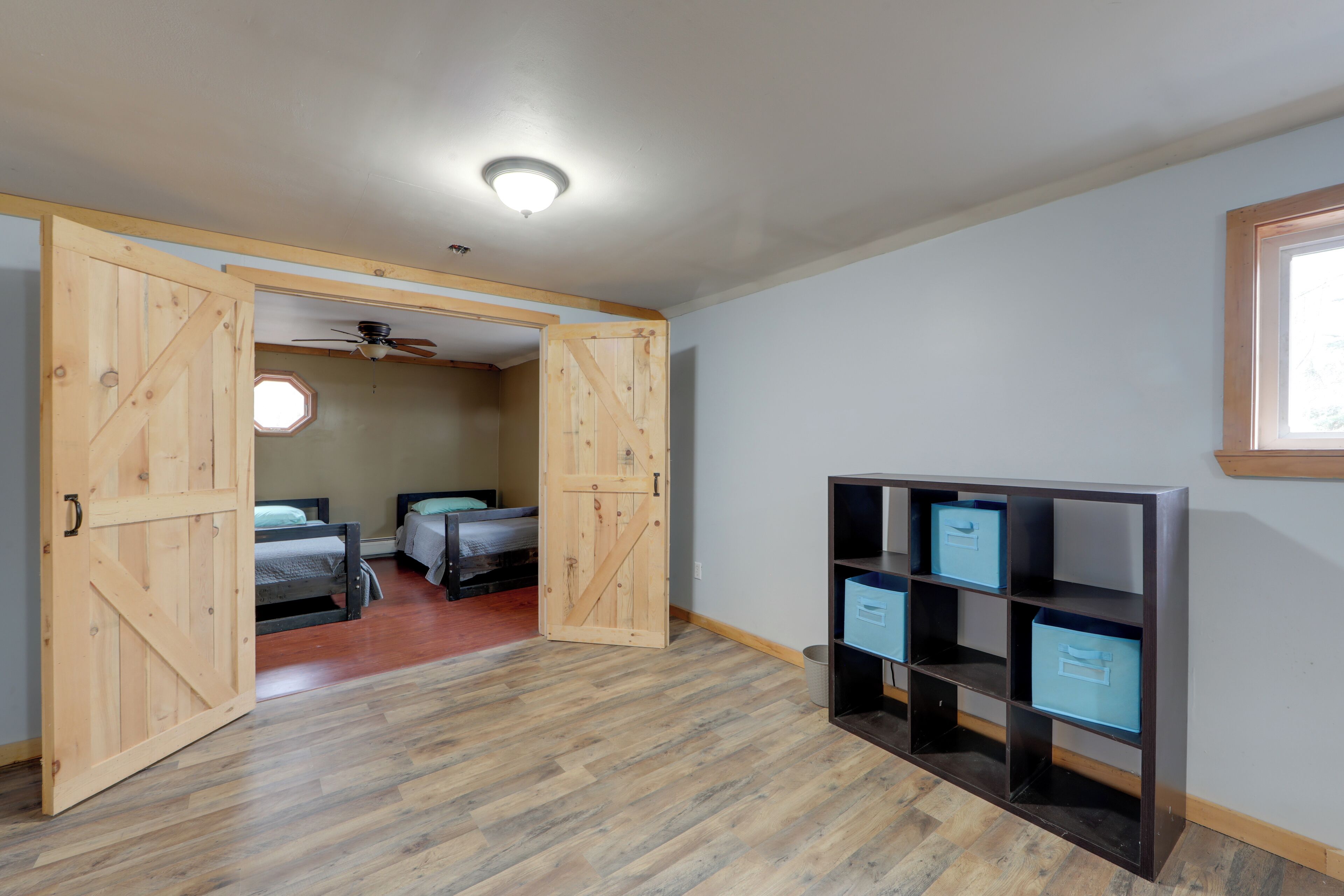 Una habitación con suelo de madera, ventilador de techo, cama con cabecera, ventana y una unidad de estanterías empotrada con cajas de almacenamiento.