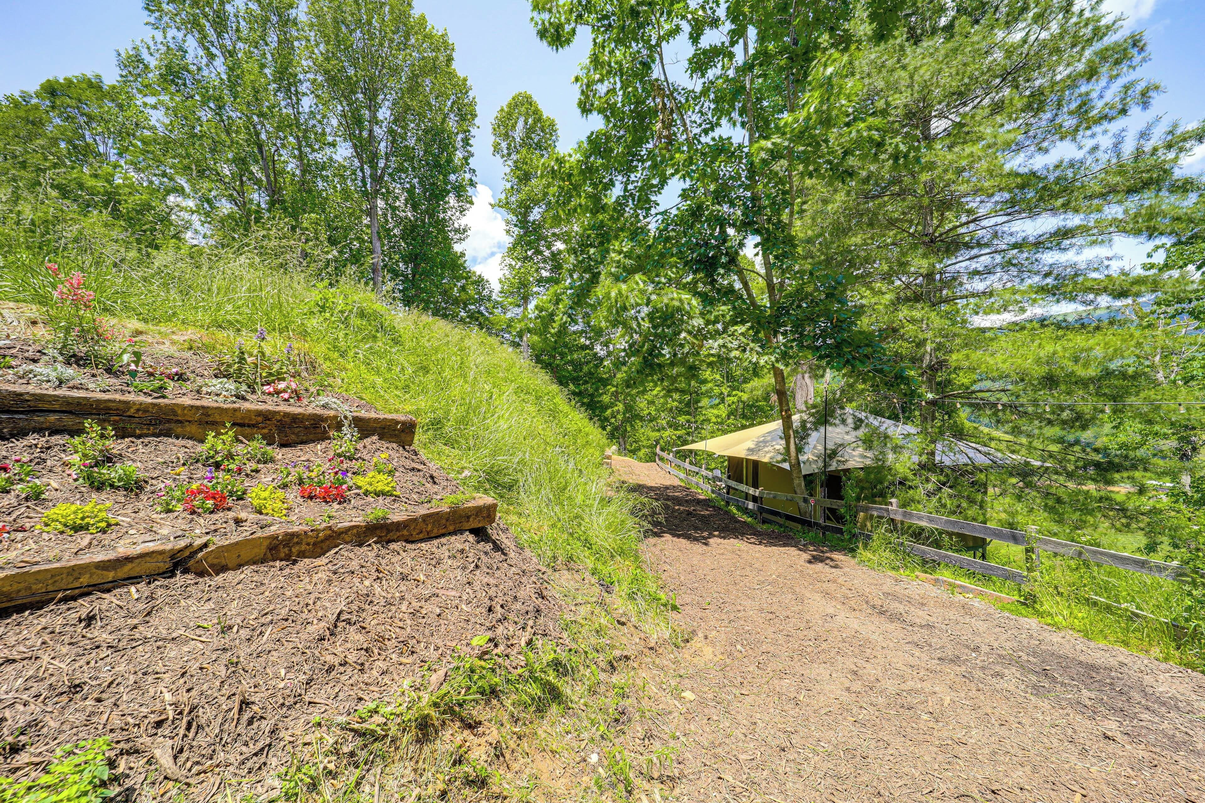 A garden with raised beds and a wooden structure, surrounded by trees and a dirt path.