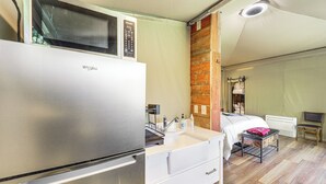 Ein kleines Zimmer mit einer Küchenzeile, inklusive Kühlschrank, Mikrowelle und Spüle. Es gibt ein Bett mit einem Holz-Kopfstück, einen kleinen Tisch und einen Stuhl.