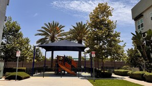 A playground with a slide and climbing structures, surrounded by palm trees and a covered play area.