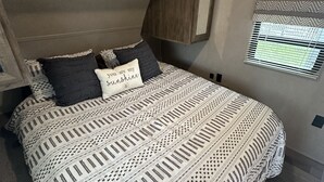 Une chambre à coucher comprenant un lit, des oreillers, une tête de lit, une fenêtre avec des stores et une étagère intégrée.