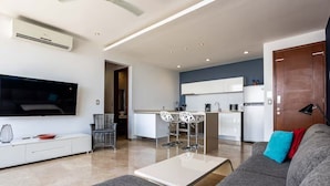 A modern living room with a grey sofa, a flat-screen TV, and a kitchen area with white cabinets and bar stools. - Beautiful 2bd-2bth - Ocean Views (Playa del Carmen)