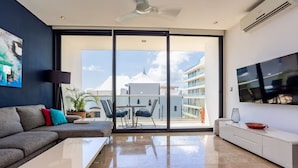 A modern living room with a grey sofa, a white coffee table, and a TV on the wall. There is a balcony with a view of buildings and a clear sky. - Beautiful 2bd-2bth - Ocean Views (Playa del Carmen)