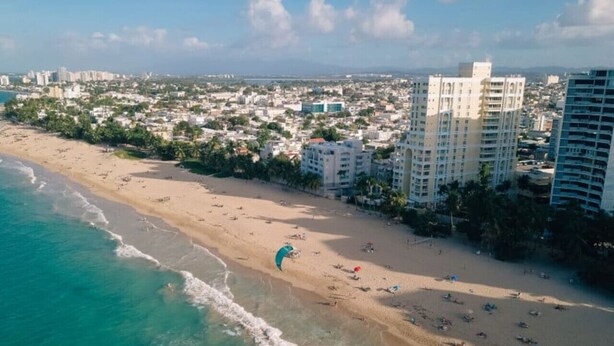 Vista aérea de una playa con paracaídas, una orilla arenosa y edificios al fondo.