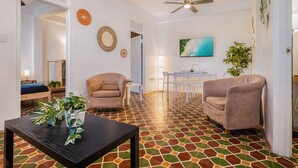 Amplia sala de estar con suelo de baldosas, dos sillones, una mesa de centro y un comedor con un televisor montado en la pared.