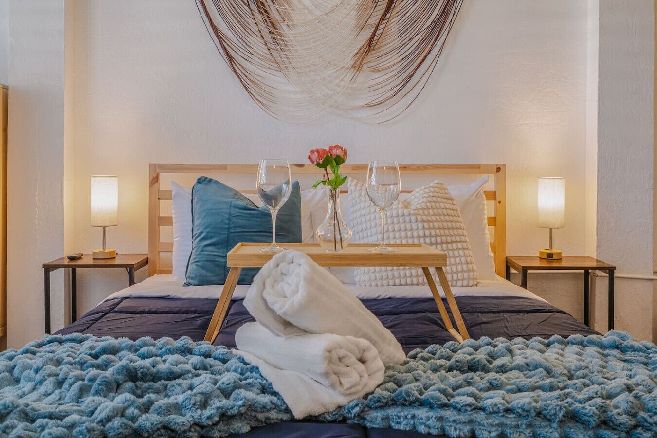 Una cama bien tendida con un edredón azul, una toalla blanca y una bandeja con un vaso de agua y un florero con flores.