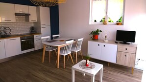 A modern kitchen with white cabinets, a wooden dining table, and chairs. There's a small white table with a plant and a decorative item. A TV is on a wooden cabinet, and a window with curtains is visible.