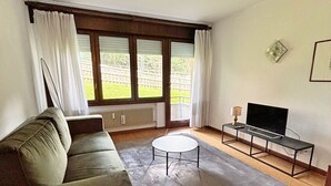 A living room with a sofa, a coffee table, a TV on a stand, and a window with curtains. - Presanella Home 4 MhD (Madonna di Campiglio)
