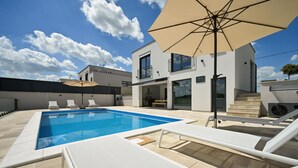 A modern pool area with lounge chairs and umbrellas, a two-story building with large windows, and a clear blue sky. - Beautiful home in Maglenca with sauna (Maglenca)