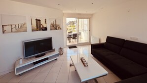 Una sala moderna con una televisión de pantalla plana, un sofá negro y una mesa de centro. Hay tres cuadros en la pared y una puerta corrediza de vidrio que da a un área exterior.