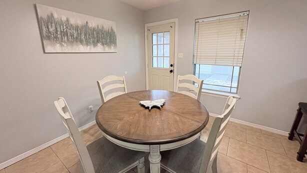 A dining room with a round table, four chairs, a painting on the wall, a door, and a window with blinds.