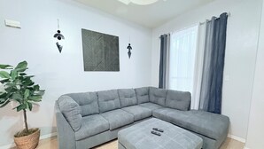 A living room with a grey sectional sofa, a matching ottoman, a potted plant, and two wall-mounted light fixtures.
