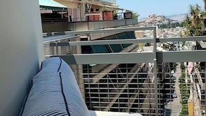 Een balkon met een gestreept kussen, een metalen hekwerk en een uitzicht op een stadsgezicht met gebouwen en een heuvel in de verte.
