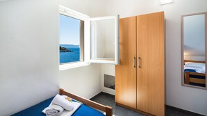 A bedroom with a wooden wardrobe, a bed with a blue mattress, a window with a view of the sea, and a small mirror on the wall.