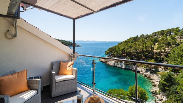 A balcony with a view of a coastal area, featuring a chair and a table with a bottle and a hat.