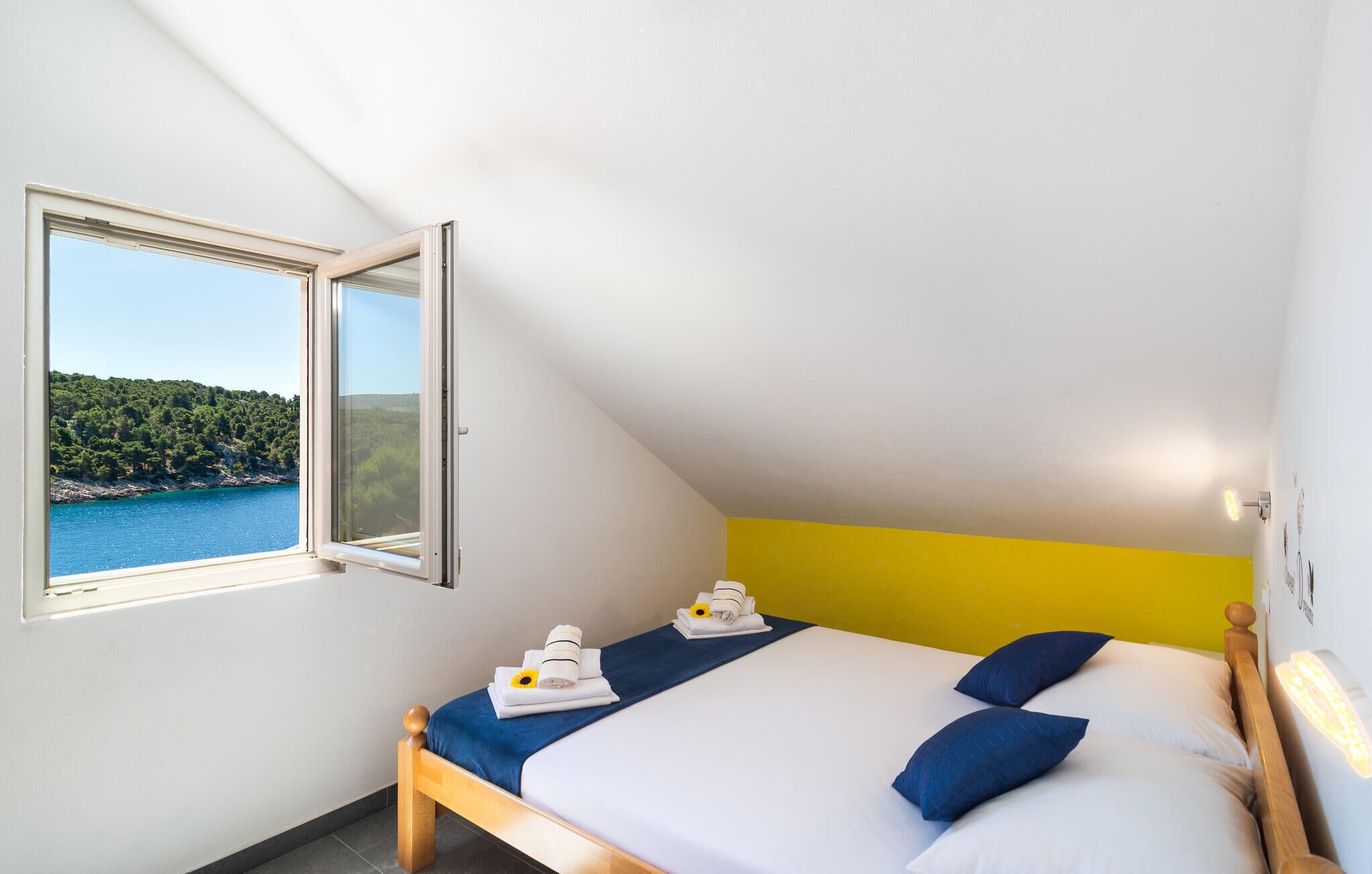 A bedroom with a bed, a window with a view of a body of water and greenery, and a yellow accent wall.