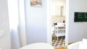 A hotel room with a white bed, a white desk, a white chair, a white bathroom with a sink and mirror, and a checkered floor. - Vatican Pixel Apartments - DOUBLE (Unknown)