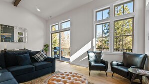 Un salon spacieux avec un canapé bleu foncé, un pouf en cuir et deux fauteuils en cuir. De grandes fenêtres offrent une vue sur les arbres et un balcon.