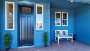 Une maison bleue avec une porte d’entrée sombre, deux plantes en pot et un banc blanc sur le porche.