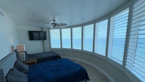 A bedroom with two beds, a ceiling fan, a TV, and large windows with blinds. - 24th Floor OceanView Oasis (Daytona Beach Shores)