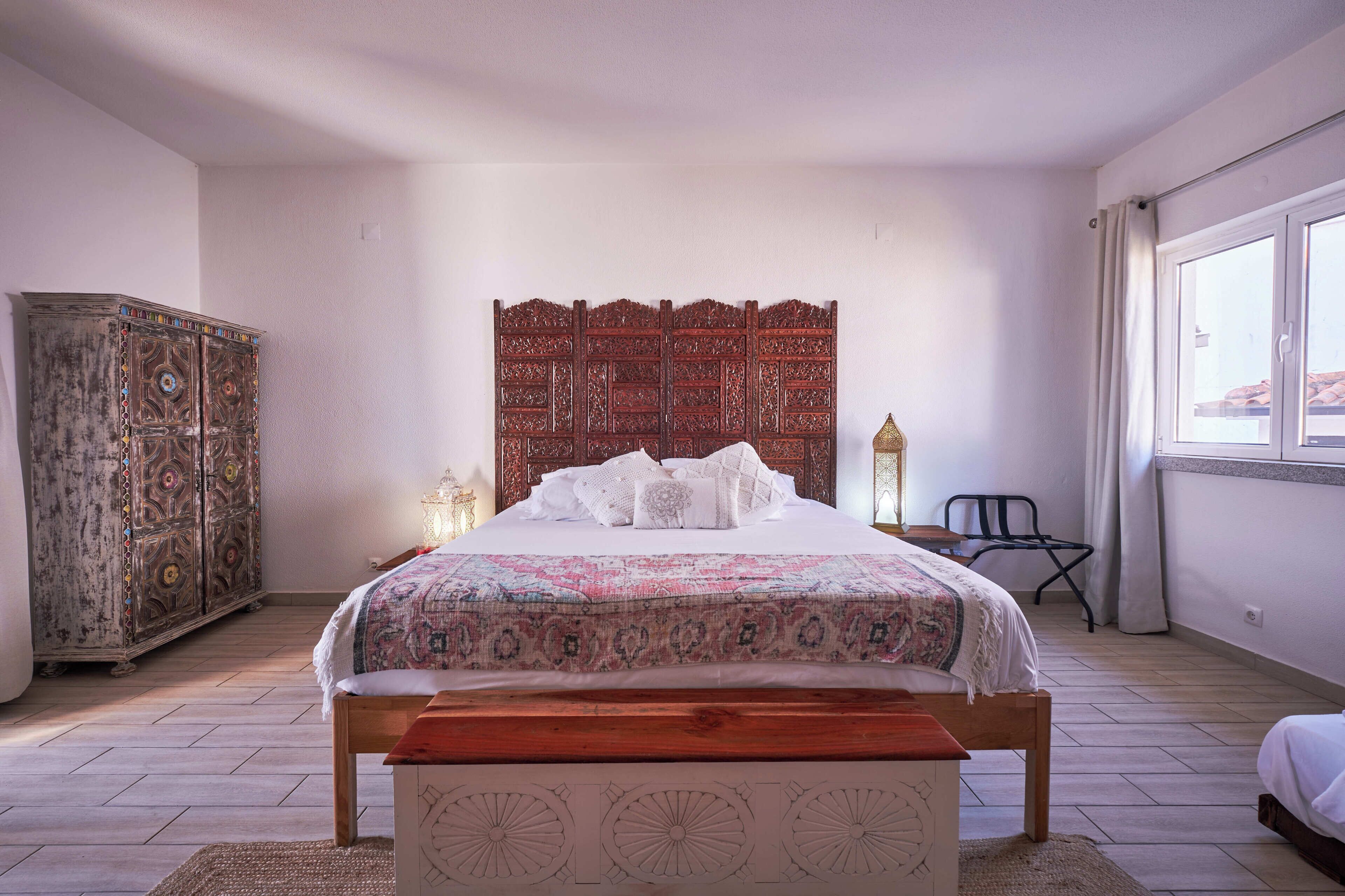 A bedroom with a wooden bed, a patterned bedspread, a decorative headboard, a vintage armoire, and a window with curtains.