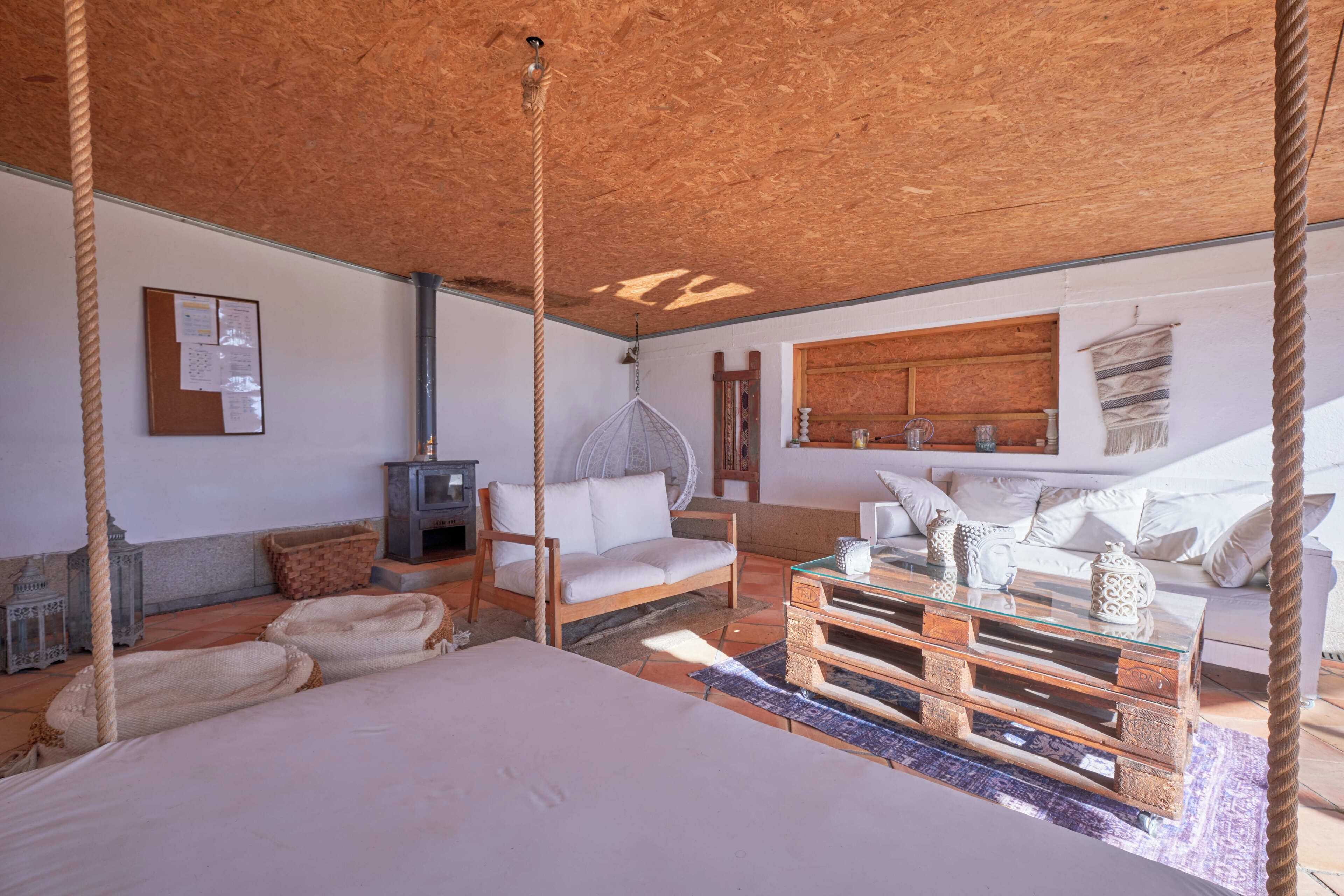 Una stanza rustica con un letto, una panca di legno, una poltrona sospesa e un tavolino da caffè in vetro.