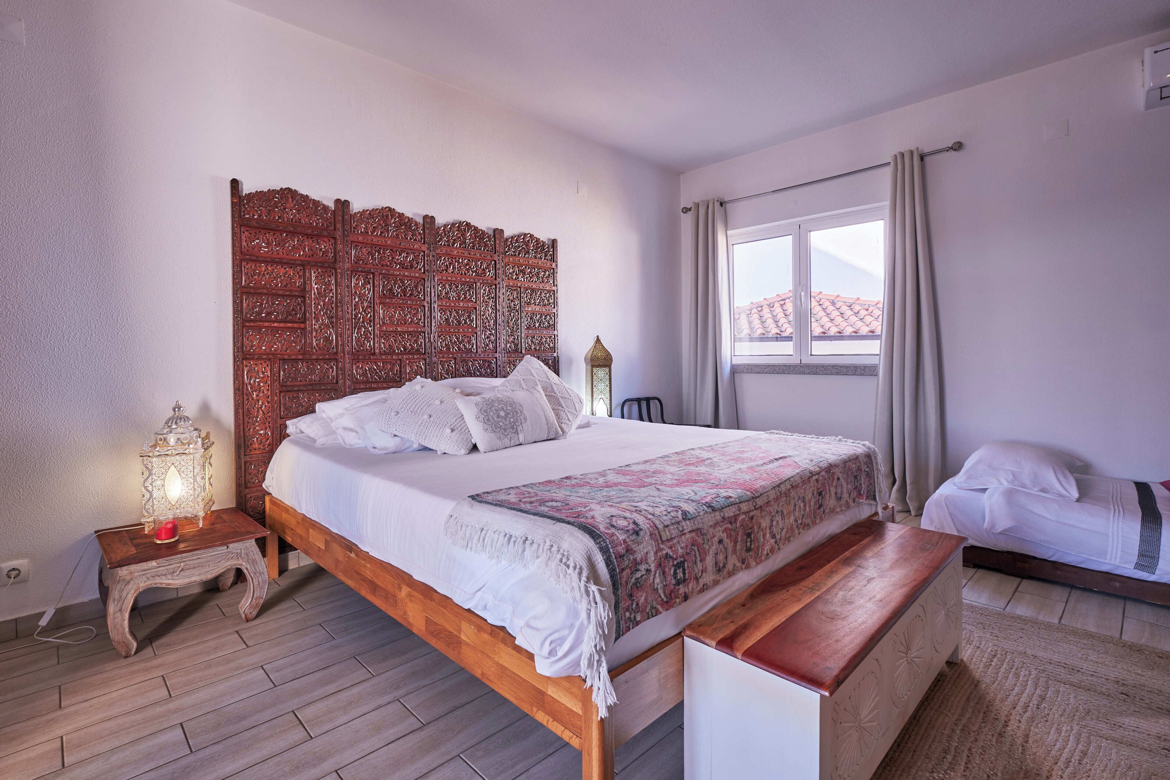 A bedroom with a wooden headboard, a bed with white linens, a nightstand with a lamp, and a window with curtains.