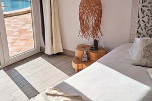 A bedroom with a large bed, a wooden nightstand with a Buddha statue, a small table with a phone and a decorative item, a window with a view of a pool, and a patterned curtain.