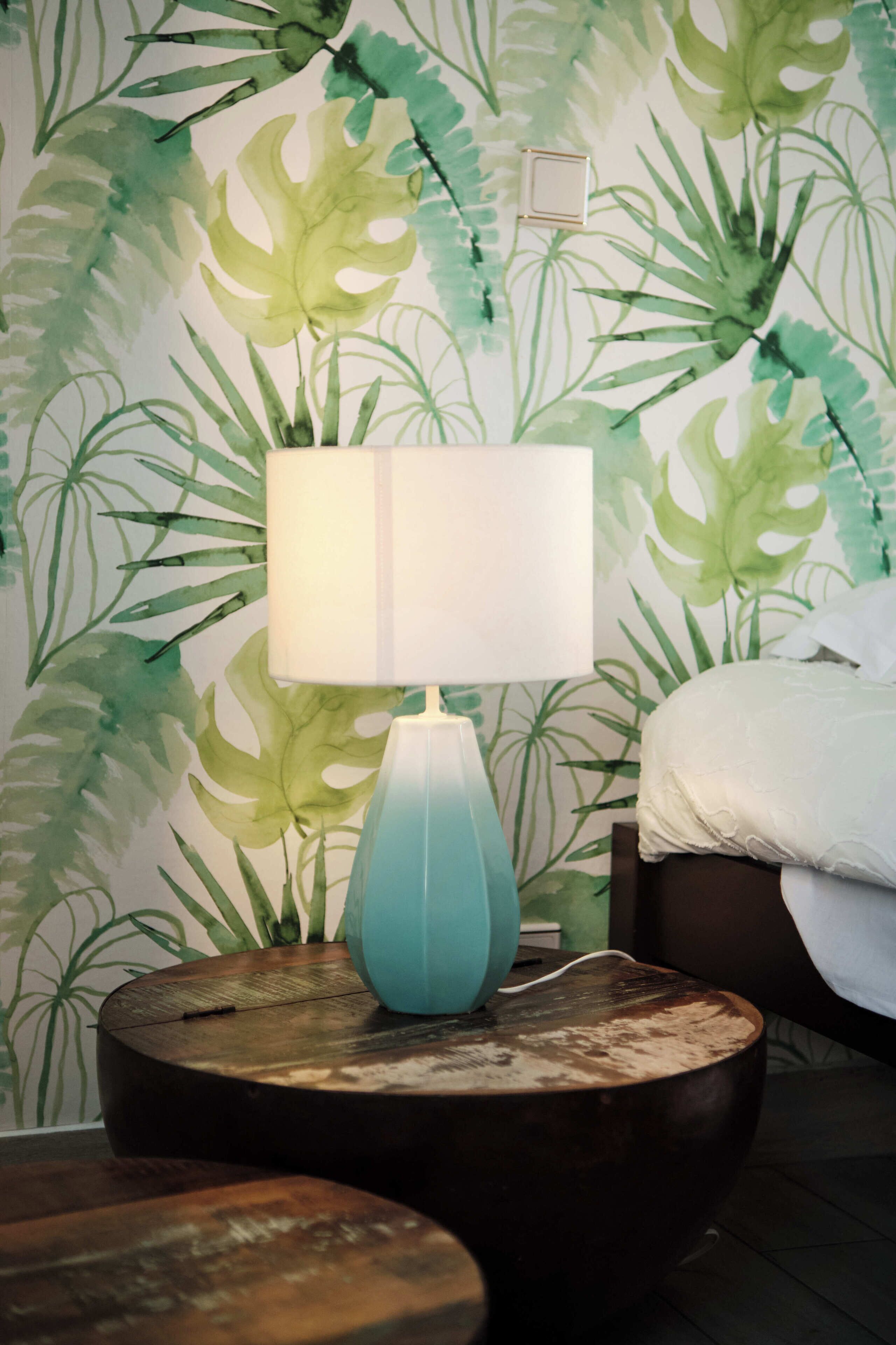 Une pièce avec une lampe turquoise posée sur un tabouret en bois, un lit recouvert de linge de lit blanc et un mur orné d’un papier peint à motifs de feuilles vertes.