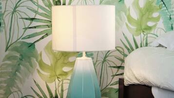 Une pièce avec une lampe turquoise posée sur un tabouret en bois, un lit recouvert de linge de lit blanc et un mur orné d’un papier peint à motifs de feuilles vertes.