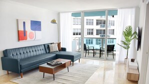 A modern living room with a sectional sofa, a coffee table, and a large window with a view of a building. - 2bd2bth Partial Ocean view Ste w/ balcony, Pool, nearby dining, beach access (Miami Beach)