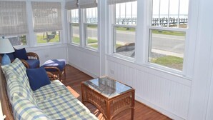 A sunlit room with a checkered sofa, a glass-top table, and a view of a beach through large windows. - Island Time- 3BR Home | Water View Chincoteague Bay (Chincoteague)