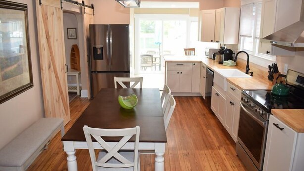 A modern kitchen with wooden flooring, white cabinetry, a stainless steel refrigerator, and a dining area with a wooden table and white chairs. - Island Time- 3BR Home | Water View Chincoteague Bay (Chincoteague)