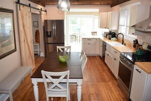 Een moderne keuken met houten vloeren, witte kasten, een roestvrijstalen koelkast en een eethoek met een houten tafel en witte stoelen.