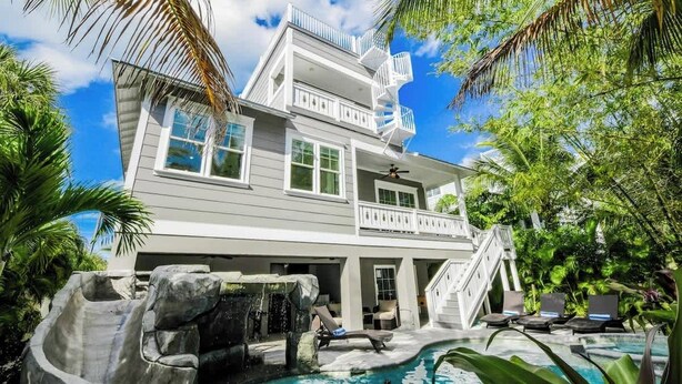 A two-story house with a swimming pool, a waterfall feature, and a balcony with a white railing. - Pineapple Paradise~WATER SLIDE/Heated Pool & Spa! (Holmes Beach)